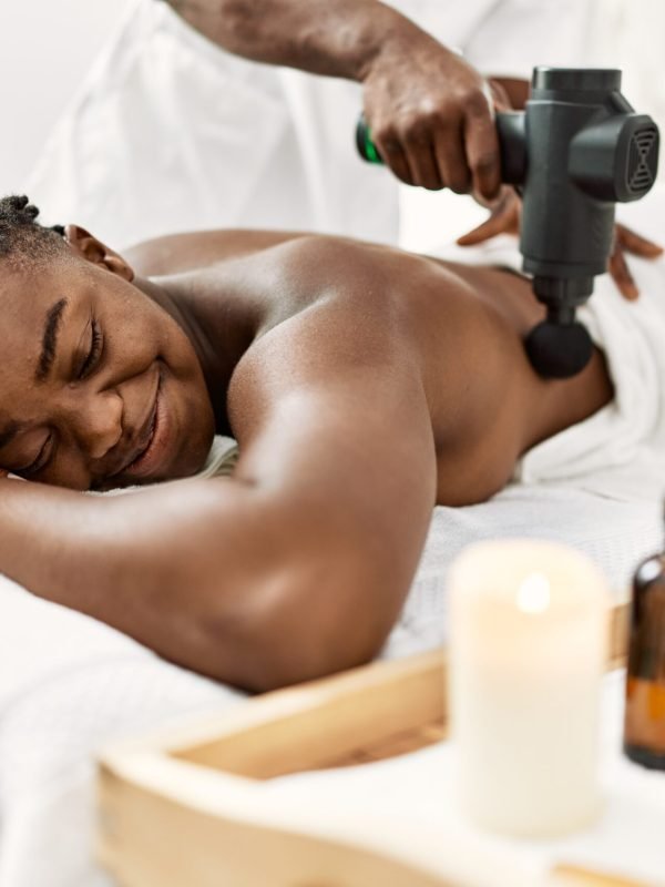 African american woman reciving back massage with gun percusion at beauty center.